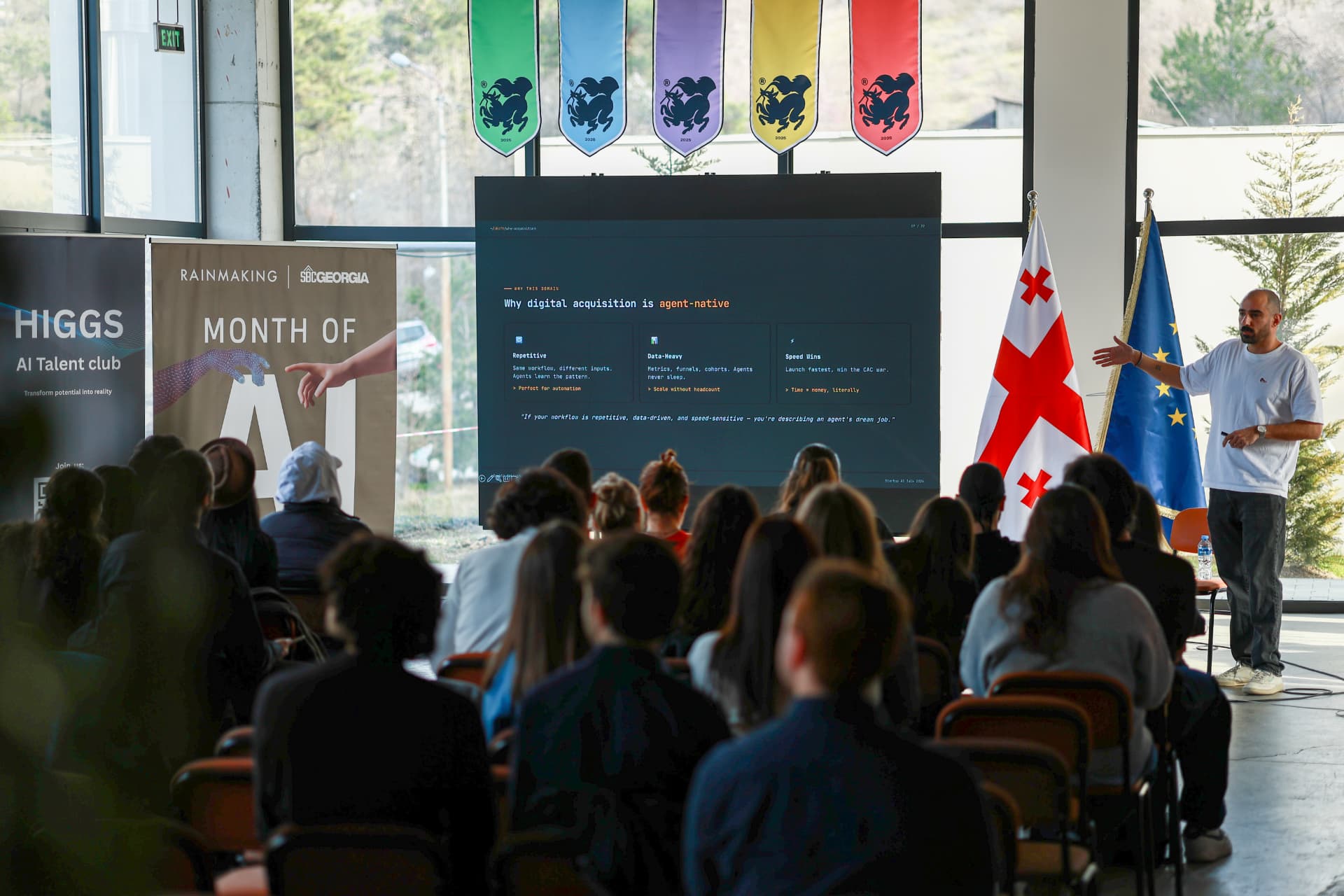 Speaker presenting to a full audience at Month of AI in Tbilisi, Georgian and EU flags beside the screen